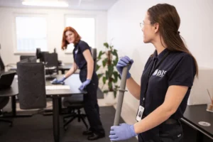 Två städare i BSIS-uniform arbetar på ett kontor, en torkar av ett skrivbord medan den andra håller en moppskaft i förgrunden.