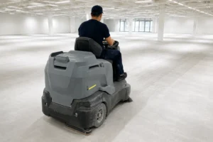 Man kör en stor golvskurmaskin genom en tom, ljus industrilokal med pelare och fönster i bakgrunden.